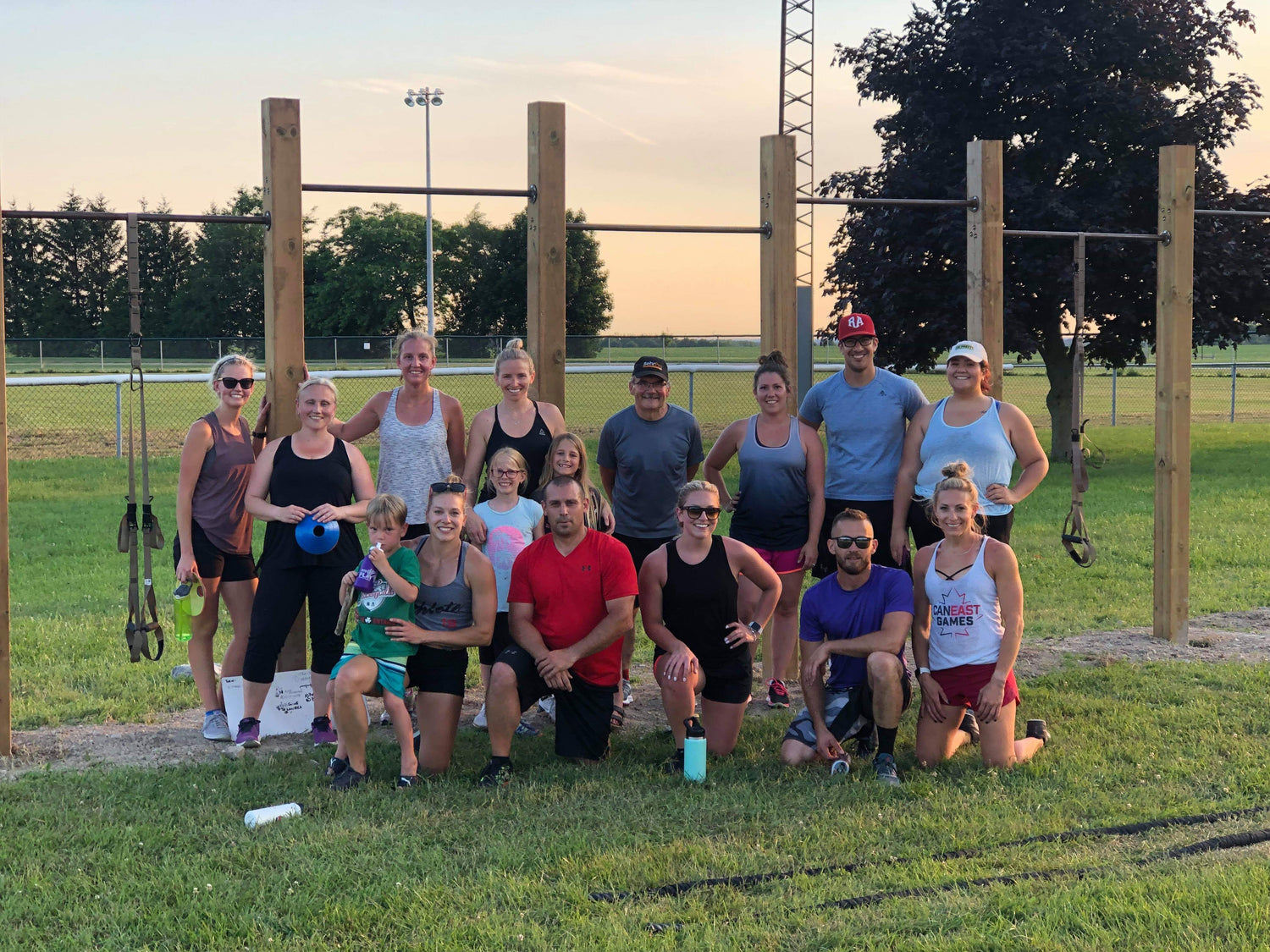Group of men, women and children in athletic wear stand in front of pull-up bars in a green field with a sunset behind them.